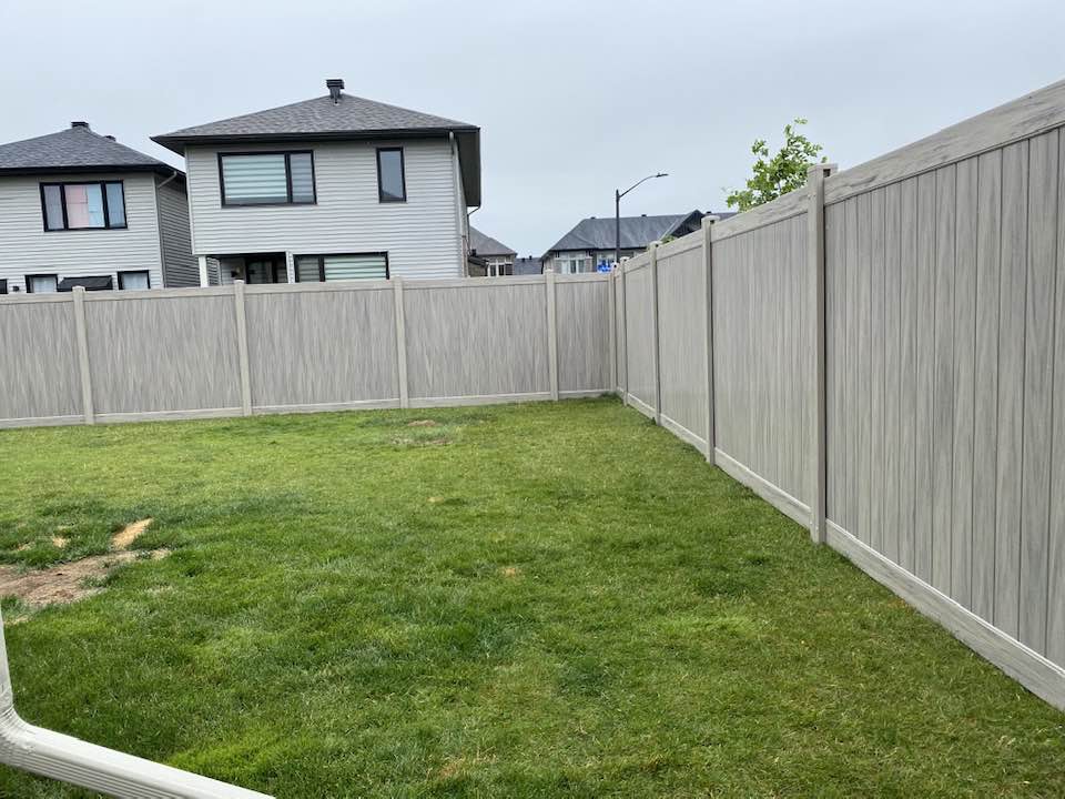 A residential backyard featuring a lush green lawn is enclosed by a beige privacy fence. Two modern houses stand in the background, framed by a gray sky.