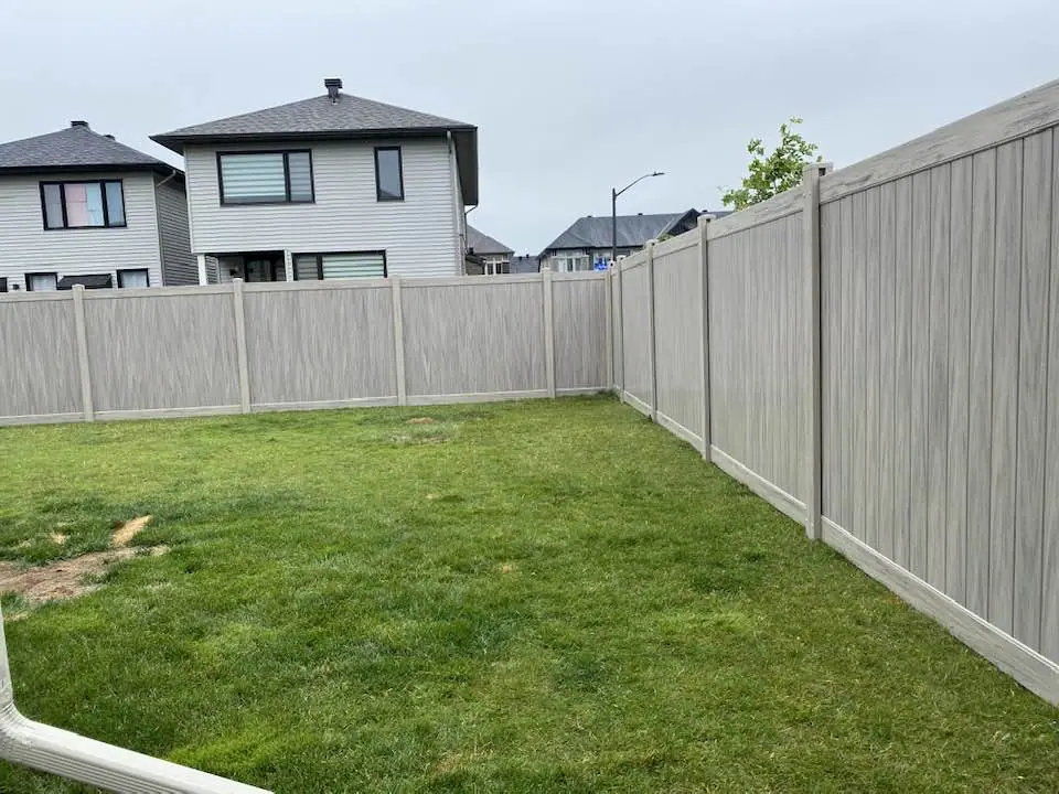 a residential backyard featuring a lush green lawn is enclosed by a beige privacy fence. two modern houses stand in the background, framed by a gray sky.