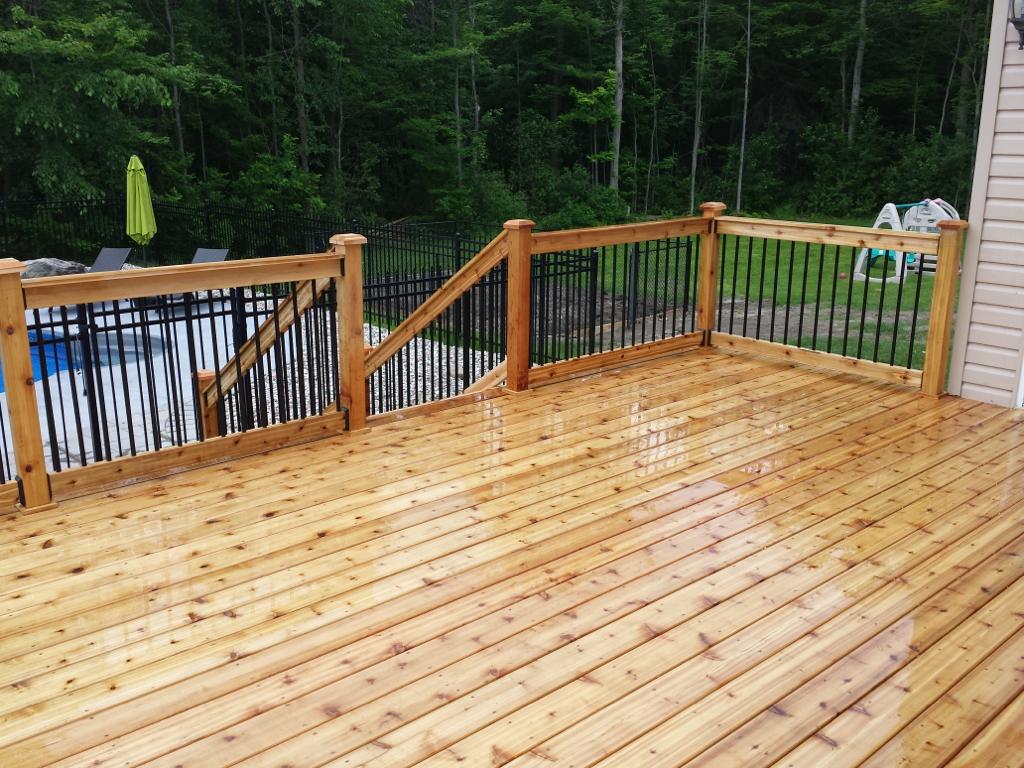 Custom wooden deck with railing, overlooking a backyard oasis featuring a pool and lawn chairs. Trees and a fence provide privacy in the background. The deck glistens, appearing wet, capturing the serene beauty after rain.