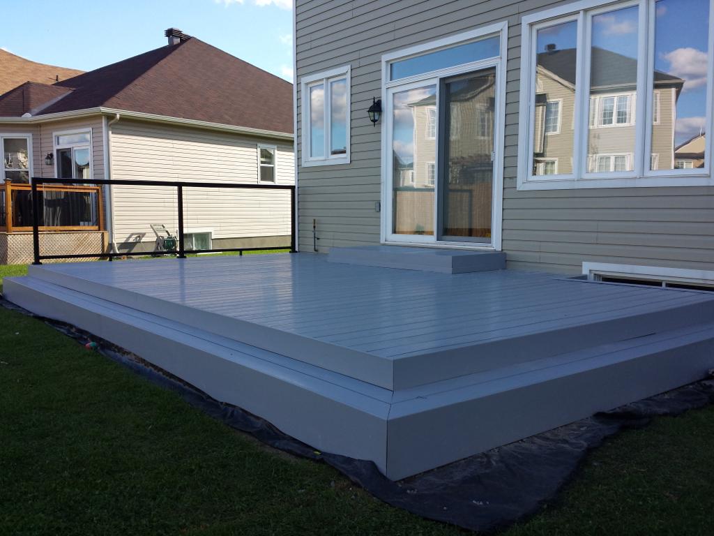 A house boasts a gray wooden deck with a step and wooden railing, epitomizing custom decks. The grassy yard surrounds it, while the neighboring house peeks through in the background.