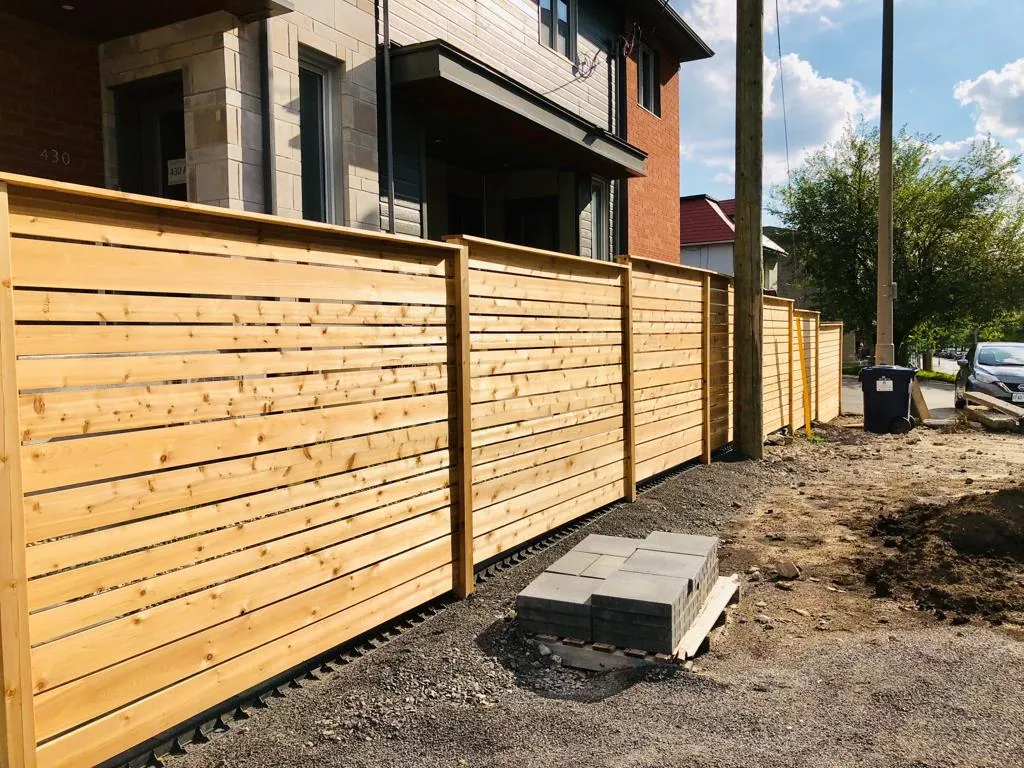 a newly constructed wooden fence lines a residential property. paving stones are stacked on the driveway, and a classic lamppost stands nearby, enhancing the charm of the setting.