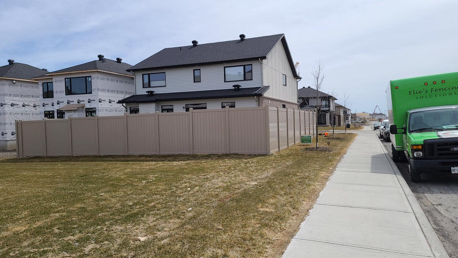 A residential street with a two-story house, beige vinyl fencing Ottawa style, a brown grass lawn, and a green company truck parked on the sidewalk.