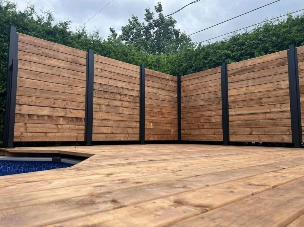 A harmonious blend of decks and fences, featuring a wooden privacy fence with horizontal planks and dark vertical supports, elegantly surrounds the pool deck, bordered by lush greenery.