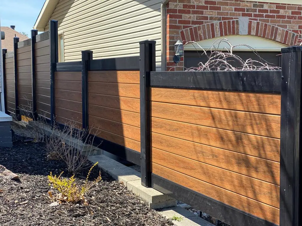A modern wood and black metal fence elegantly decorates the front of a brick house, with a small plant in the foreground and a garage door partially visible.