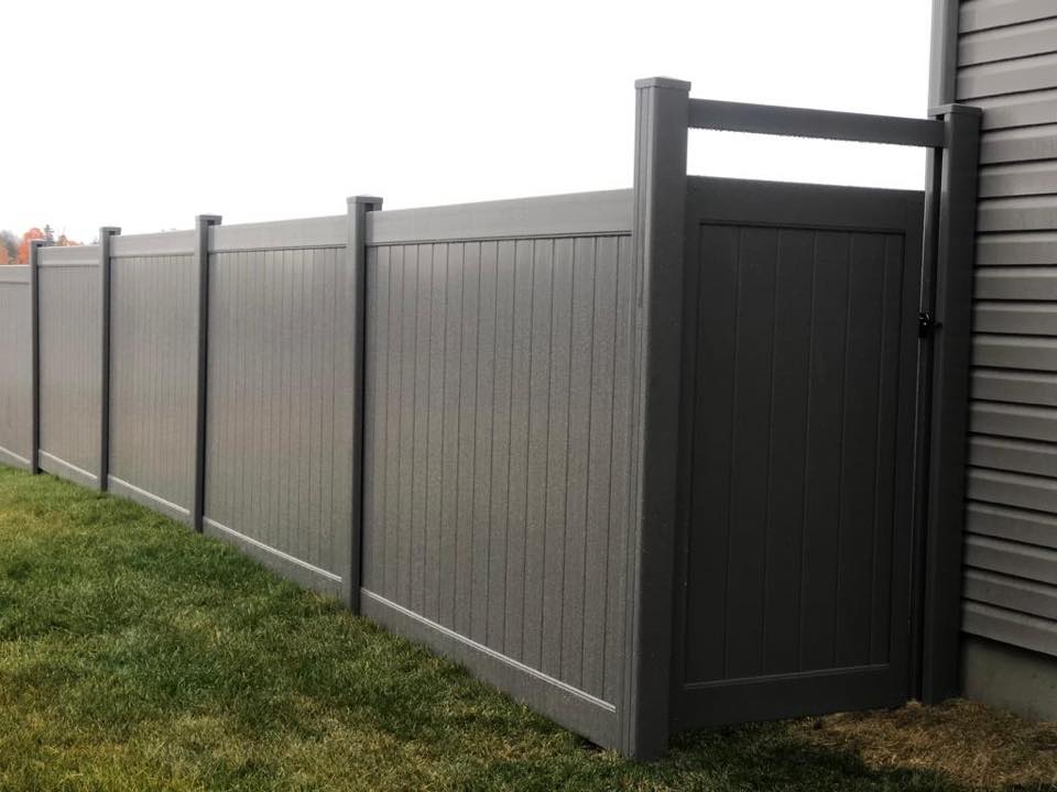 Gray vinyl fences with vertical paneling feature a tall gate, perfectly situated on a grassy lawn near a building.