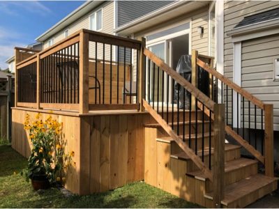 a wooden deck with a staircase and black metal railing, situated next to a residential building, with some yellow flowers in the foreground, deck design