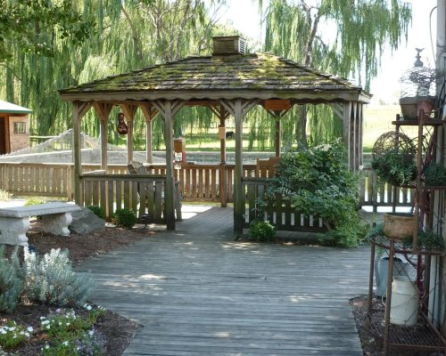 a wooden gazebo with a mossy roof, featuring gazebo construction ottawa craftsmanship, is surrounded by lush plants and trees. a stone bench rests on the left while a potted plant rack adorns the right. a wooden path winds gracefully to this tranquil spot.