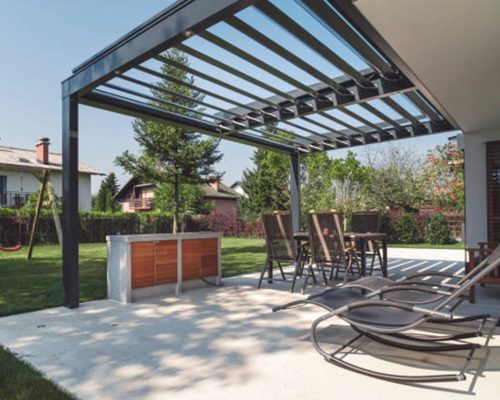 discover a modern outdoor patio designed by pergola builders barrhaven, featuring sleek bar stools, cozy lounge chairs, and lush greenery. enjoy the sunny day with two charming houses visible in the background.